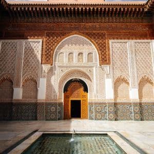 Un patio de piedra con una piscina central frente a un edificio ornamentado con puertas de madera talladas y azulejos intrincados.