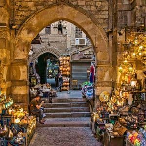 Une allée en pierre dans un marché animé est remplie d'étals présentant de nombreuses lanternes colorées en verre et en métal illuminées.