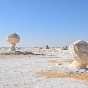 De grandes formations rocheuses de craie blanche se dressent dans un paysage désertique de sable blanc et jaune sous un ciel bleu clair.