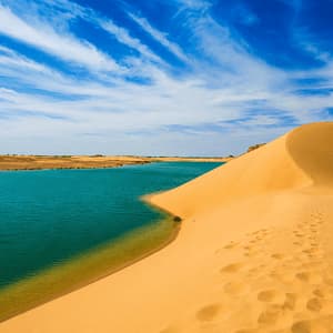 Une grande dune de sable doré avec des empreintes de pas rencontre un plan d'eau turquoise dans le désert sous un ciel bleu avec des nuages.