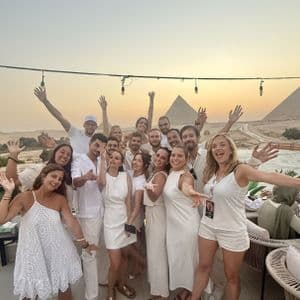 Un groupe WeRoad, habillé de blanc, pose sur une terrasse avec les pyramides de Gizeh en arrière-plan, au coucher du soleil.
