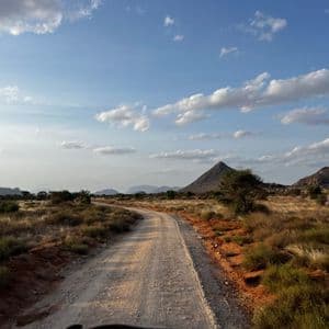 A winding dirt road through a dry savanna landscape, with distant mountains under a blue sky with scattered clouds.