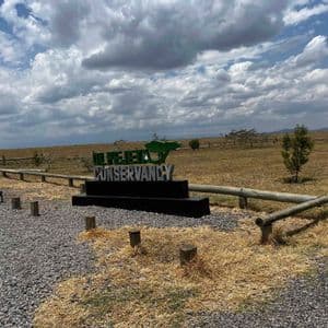 The Ol Pejeta Conservancy entrance sign stands in a dry, grassy field bordered by a wooden fence under a cloudy sky.