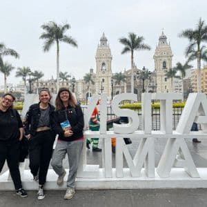 Tre donne in viaggio di gruppo WeRoad sorridono e posano accanto a un grande cartello bianco "Visita Lima" in una piazza cittadina con una cattedrale sullo sfondo.