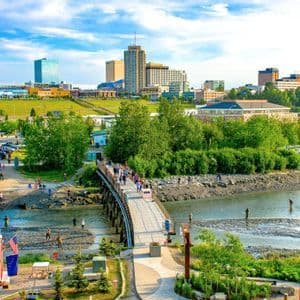 La gente attraversa una passerella di legno su un fiume, mentre altri pescano nell'acqua sottostante, con lo skyline di una città sullo sfondo.