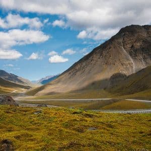 Una strada tortuosa attraversa una vasta valle di montagna coperta di tundra verde e gialla sotto un cielo blu con nuvole bianche.