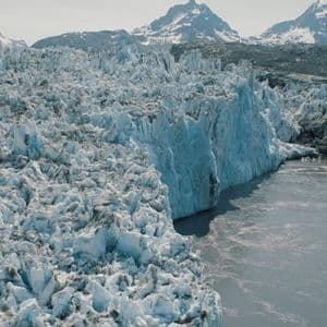 L'estremità di un enorme ghiacciaio dalla superficie irregolare incontra uno specchio d'acqua di fronte a una catena di montagne innevate.