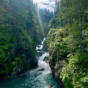 Una cascata a più livelli si getta da una gola ripida e boscosa in un fiume turchese e vibrante sottostante, con un raggio di sole che filtra tra gli alberi.