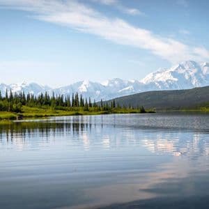 Un lago sereno riflette una fila di alberi sempreverdi e una catena montuosa innevata in lontananza sotto un cielo azzurro.