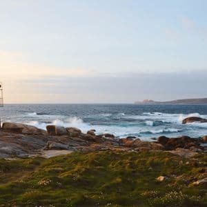 Un faro se alza en una costa rocosa mientras las olas rompen en la orilla, con una zona de césped en primer plano.