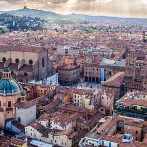 Vista aerea di una città storica con tetti di tegole rosse, una grande basilica, una cupola verde e una piazza pubblica, con colline sullo sfondo.