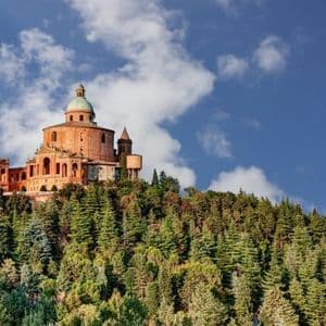 Una grande basilica color arancio con una cupola verde si erge su una collina ricoperta da fitti alberi verdi sotto un cielo parzialmente nuvoloso.