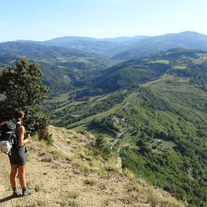 Un escursionista con un grande zaino è su un sentiero di montagna, che osserva un paesaggio verde e collinare.