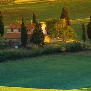Un casale in pietra con tetto in tegole è circondato da cipressi su una dolce collina verdeggiante durante l'ora d'oro.