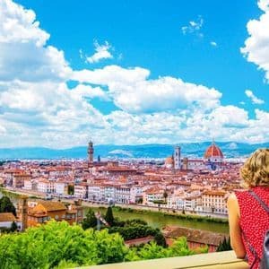 Una donna con uno zaino osserva da un punto panoramico una città storica con una grande cupola e un fiume sotto un cielo blu nuvoloso.