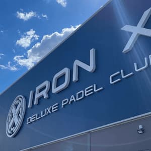 Inquadratura dal basso dell'insegna 'Iron X Deluxe Padel Club' con scritte bianche su un edificio blu, sotto un cielo parzialmente nuvoloso.