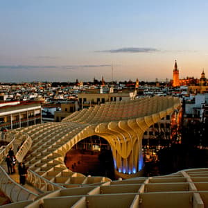 Una vista dal tortuoso camminamento di una grande e moderna struttura in legno che si affaccia su un paesaggio urbano con una cattedrale illuminata al tramonto.