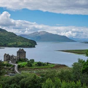 Un château de pierre sur une île dans un loch, relié par un pont à la rive, avec des montagnes en arrière-plan sous un ciel partiellement nuageux.
