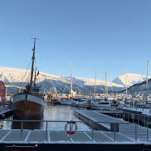 A traditional wooden boat is docked in a snowy harbor with mountains in the background under a clear, sunny sky.