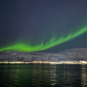 A green aurora borealis glows in the starry night sky above snowy mountains and a town reflecting on the water.