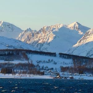 A small village on a snowy coast with blue water in the foreground and a large, sunlit mountain range behind.