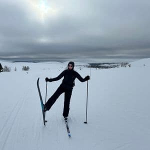 A person in black ski gear poses playfully on cross-country skis in a vast, snowy landscape under an overcast sky.