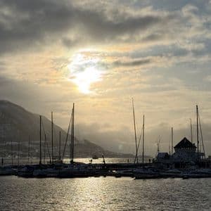 Sailboats docked in a snowy harbor at sunset, with light reflecting on the water and a mountain in the background.
