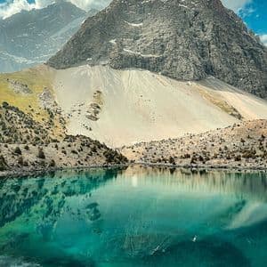 Un'imponente montagna rocciosa e un cielo nuvoloso si riflettono nelle acque immobili e turchesi di un lago alpino.
