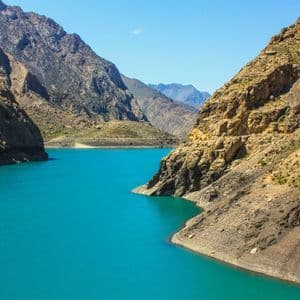 Un vivace lago turchese scorre attraverso un profondo canyon, circondato da ripide montagne rocciose sotto un cielo azzurro e limpido.