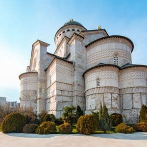 Una grande cattedrale di pietra bianca con una cupola centrale e intagli intricati, circondata da un giardino verde sotto un cielo azzurro e limpido.