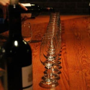 Una persona versa vino rosso da una bottiglia in una lunga fila di bicchieri vuoti preparati per una degustazione di vino su un tavolo di legno.