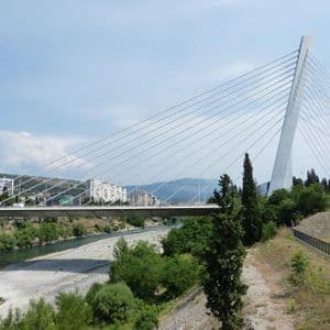 Un moderno ponte strallato bianco che attraversa un fiume poco profondo, con uno skyline cittadino e colline lontane sullo sfondo.