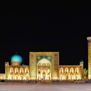 Un ornato complesso architettonico con cupola turchese e minareti è illuminato da luci dorate in un'ampia piazza di notte.