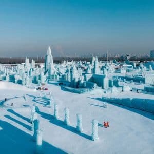 Vista aérea de un festival de invierno con grandes y complejas esculturas de hielo y nieve que cubren un paisaje nevado bajo un cielo azul despejado.
