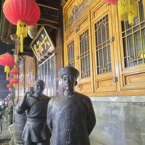 Due statue di bronzo raffiguranti uomini in abiti tradizionali si ergono di notte all'esterno di un edificio decorato con lanterne rosse appese.