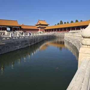 Un foso curvo con muros de piedra refleja edificios tradicionales chinos con techos de tejas naranjas bajo un cielo despejado.