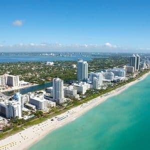 Vista aerea di una città con alti edifici bianchi, una spiaggia sabbiosa che si unisce all'oceano turchese sotto un cielo azzurro.