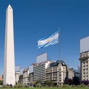 La bandiera argentina sventola accanto a un alto obelisco bianco in una piazza cittadina, con edifici storici sotto un cielo azzurro chiaro.
