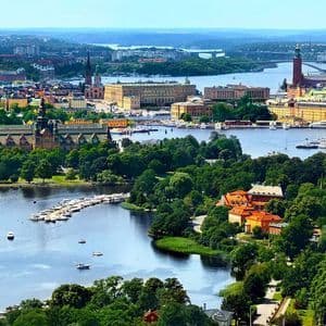 Una vista aerea di una città tentacolare lungo un corso d'acqua, con edifici storici, alberi verdi e numerose barche in una giornata limpida.