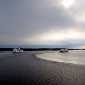 Due grandi traghetti navigano sul mare, le loro scie dividono la superficie dell'acqua, con una costa boscosa sotto un cielo nuvoloso.