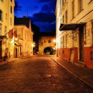 Una strada acciottolata vuota illuminata di notte, affiancata da vecchi edifici e che conduce a un arco in pietra sotto un cielo blu scuro.
