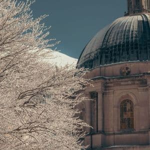 Un edificio classico con cupola si erge accanto a rami innevati, con una montagna imbiancata sullo sfondo.