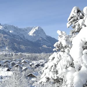 Ein schneebedecktes Dorf liegt in einem Bergtal unter strahlend blauem Himmel, der Blick geht vorbei an stark schneebeladenen Bäumen.