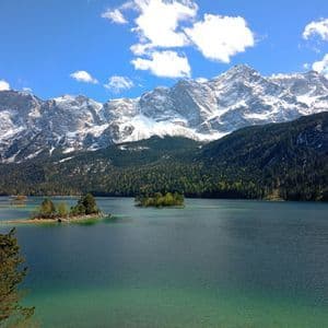 Ein türkiser See mit kleinen, bewaldeten Inseln liegt am Fuße einer schneebedeckten Bergkette unter blauem Himmel mit Wolken.