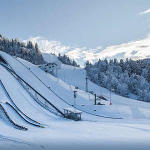 Eine große Skisprunganlage mit mehreren Schanzen an einem schneebedeckten Berghang, umgeben von Pinien.