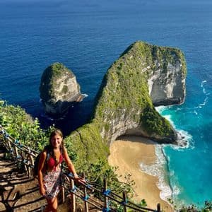 Eine Frau steht auf einem Klippenweg und blickt auf eine einzigartige, grün bewachsene Felsformation und einen Sandstrand mit türkisfarbenem Wasser.