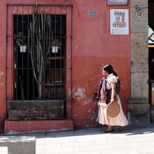 Una mujer con traje tradicional y mascarilla camina por una acera junto a un edificio rojo-naranja con una ventana enrejada.