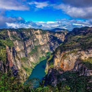 Un río turquesa fluye por un cañón profundo con acantilados empinados y cubiertos de vegetación bajo un cielo azul con nubes dispersas.