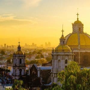A historic cathedral with golden domes overlooking a city with a modern skyline during a golden sunset.