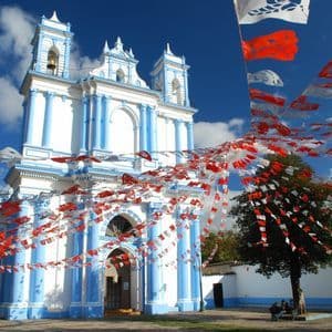 Eine weiß-blaue Kirche ist mit rot-weißen Papierfahnenketten auf einem Platz unter einem klaren blauen Himmel geschmückt.